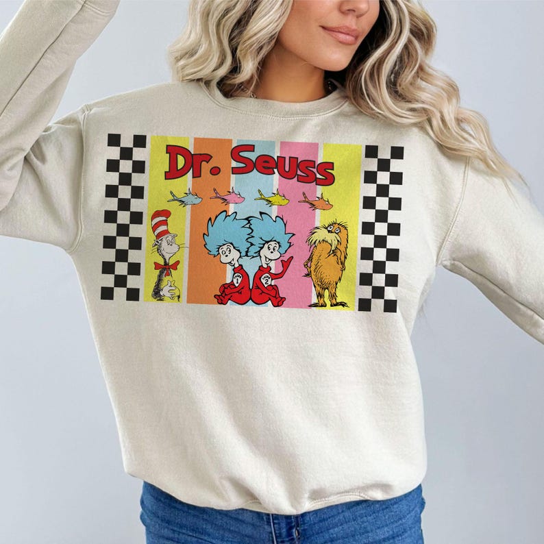 Puede incluir: Sudadera color crema con un colorido gr&aacute;fico de Dr. Seuss. El gr&aacute;fico incluye el texto "Dr. Seuss" en rojo, junto con ilustraciones de personajes como el Gato en el Sombrero, los Cosas y el Lorax. Patrones de cuadros blancos y negros en los lados.