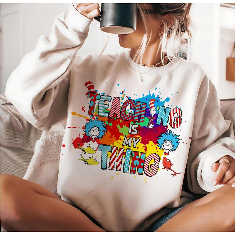 May include: Cream-colored sweatshirt with the phrase "TEACHING IS MY THING" in colorful, playful lettering. The design includes cartoon characters and paint splatter effects, creating a vibrant and whimsical look. A fun and stylish apparel item.