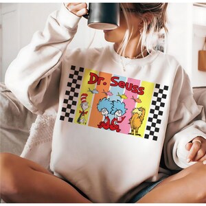 Puede incluir: Sudadera de color crema con un colorido dise&ntilde;o de Dr. Seuss. El dise&ntilde;o incluye personajes como el Gato en el Sombrero, Thing 1 y Thing 2, y el Lorax, con las palabras "Dr. Seuss" en negrita. Patrones de cuadros enmarcan el dise&ntilde;o.