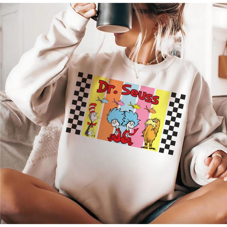 Puede incluir: Sudadera de color crema con un colorido dise&ntilde;o de Dr. Seuss. El dise&ntilde;o incluye personajes como el Gato en el Sombrero, Thing 1 y Thing 2, y el Lorax, con las palabras "Dr. Seuss" en negrita. Patrones de cuadros enmarcan el dise&ntilde;o.