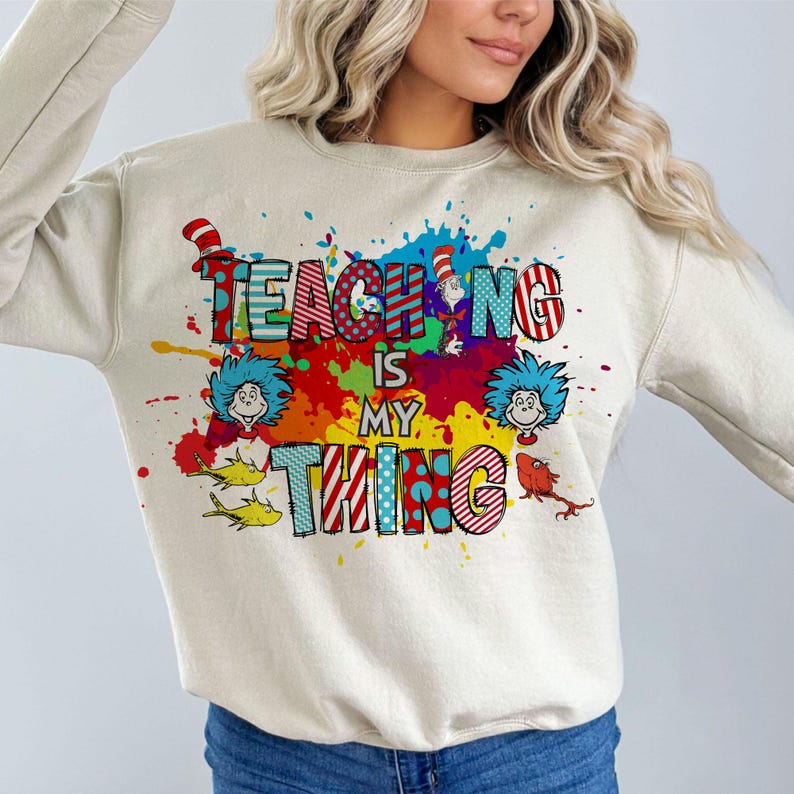 May include: A beige sweatshirt with the text "TEACHING IS MY THING" in colorful, patterned letters. The design includes cartoon characters and paint splatters in red, blue, yellow, and green.