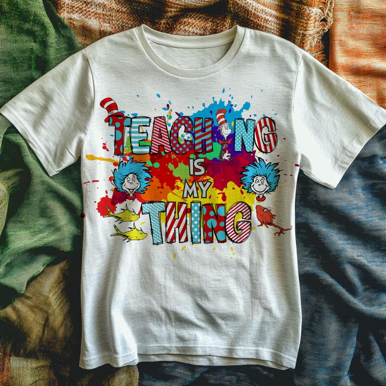 May include: White t-shirt with the text "TEACHING IS MY THING" in colorful, patterned letters. The design includes cartoon characters and splashes of red, blue, yellow, and green. A fun and vibrant graphic tee.