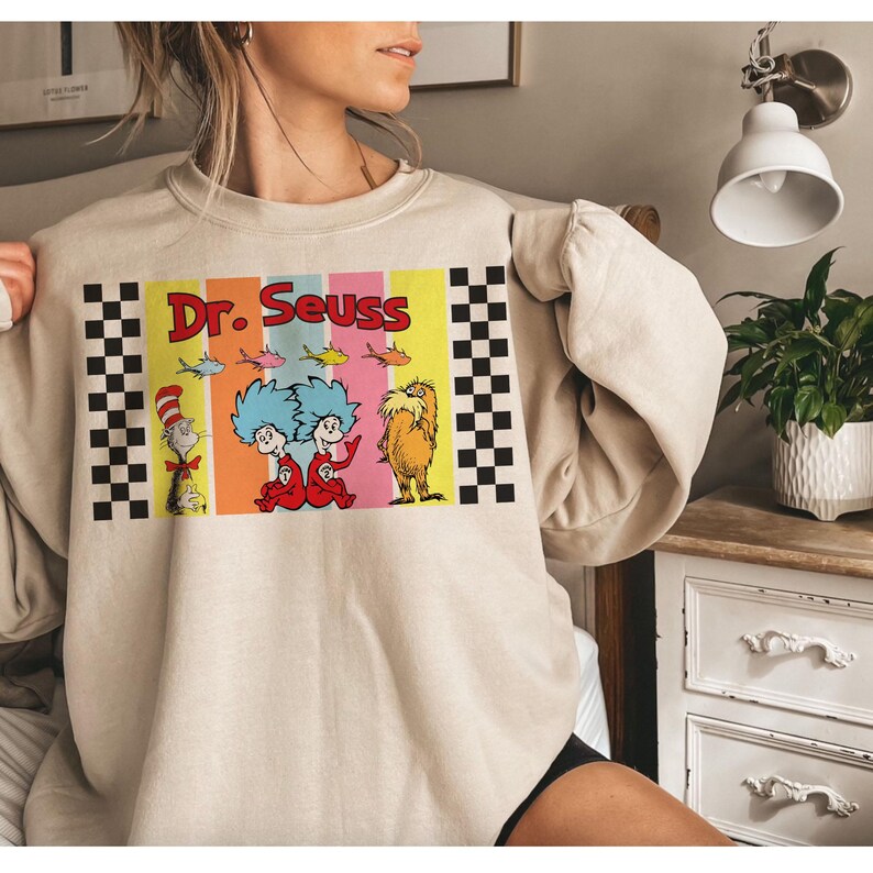 Puede incluir: Sudadera beige con un gr&aacute;fico colorido de Dr. Seuss. El gr&aacute;fico incluye el texto "Dr. Seuss" e ilustraciones de personajes como el Gato en el Sombrero, Thing 1 y Thing 2, y el Lorax. Patrones de cuadros enmarcan el gr&aacute;fico.