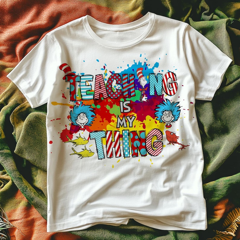 May include: White t-shirt with the phrase "TEACHING IS MY THING" in colorful, patterned letters. The design includes illustrations of characters and splashes of paint in various colors, creating a playful and vibrant look.