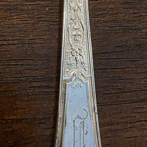 May include: Silver butter knife with a wide, rounded blade and a decorative handle. The handle features ornate floral and scrollwork designs, and an initial is engraved at the base. The knife is set against a dark background.