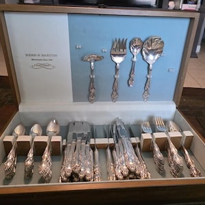 May include: A wooden silverware chest with a light blue interior, showcasing a collection of Reed & Barton silverware. The set includes forks, spoons, knives, and serving utensils, all with ornate, detailed handles.