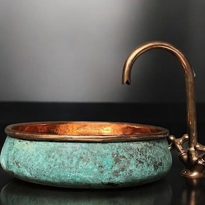 May include: A copper sink with a verdigris finish and a matching faucet. The sink has a rounded shape with a copper rim. The faucet has a curved spout and cross handles. The sink and faucet are set against a dark background.