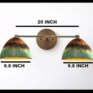May include: A wall-mounted light fixture with two dome-shaped shades. The shades are green, brown, and orange. The fixture is 20 inches wide and each shade is 9.8 inches wide. The fixture has a bronze finish.