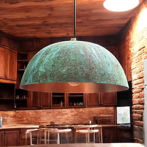 Puede incluir: Una gran lámpara colgante en forma de cúpula con un acabado de cobre y una pátina verde. La luz cuelga de un cable negro en una cocina con gabinetes de madera y paredes de ladrillo.
