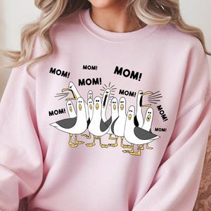 Peut inclure: Un sweat-shirt rose clair avec un motif de dessin anim&eacute; de mouettes criant le mot "MOM!". Les mouettes sont en noir et blanc avec des pieds et des becs jaunes. Le sweat-shirt est de style col rond.