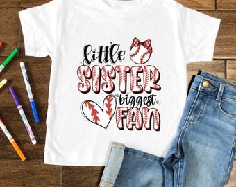 May include: A white t-shirt with the text "Little Sister Biggest Fan" in black and red, with baseball-themed graphics. The shirt is paired with blue denim jeans. Colorful markers are scattered on a wooden surface.