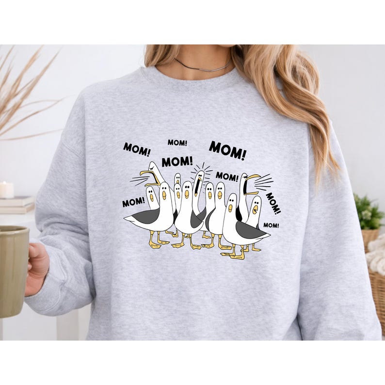 Peut inclure: Un sweat-shirt gris clair avec un motif de dessin anim&eacute; de mouettes. Les mouettes sont noires, blanches et jaunes, avec le mot "MOM!" dans des bulles. Le sweat-shirt est de style d&eacute;contract&eacute;.