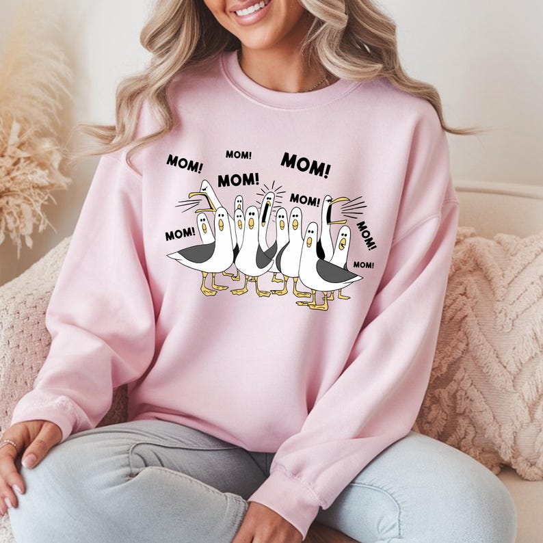Peut inclure: Un sweat-shirt rose clair avec un motif de dessin anim&eacute; de mouettes criant le mot "MOM!". Les mouettes sont en noir et blanc avec des pieds et des becs jaunes. Le sweat-shirt est de style col rond.