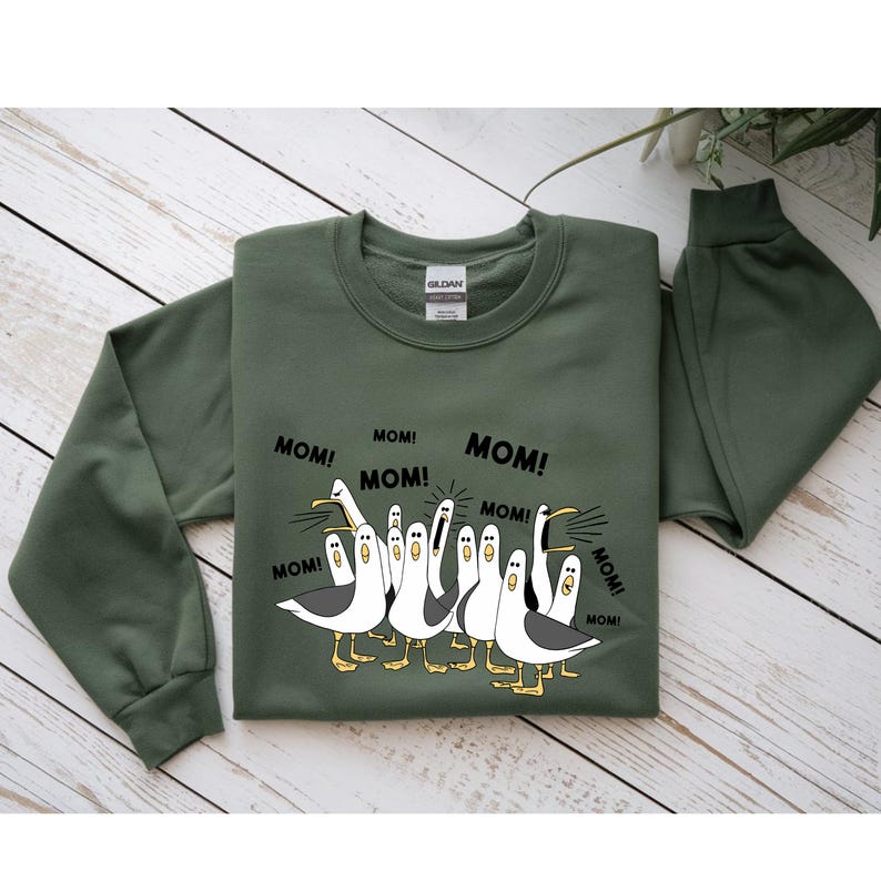 Peut inclure: Un sweat-shirt vert olive avec une illustration de mouettes criant le mot "MOM!". Les mouettes sont blanches avec des d&eacute;tails noirs et des pieds jaunes. Le sweat-shirt a un col rond et des manches longues.
