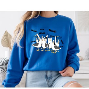 Peut inclure: Sweat-shirt bleu roi avec une illustration de dessin anim&eacute; de mouettes. Les mouettes sont blanches avec des ailes grises et des pieds jaunes, et elles crient toutes "MOM!". Le texte "MOM!" est r&eacute;p&eacute;t&eacute; autour des mouettes.