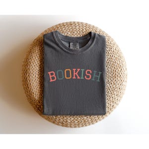 A gray t-shirt with the word "BOOKISH" printed on it, laid out on a woven basket or round seat.