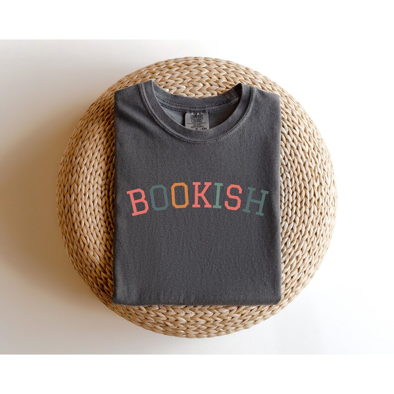 A gray t-shirt with the word "BOOKISH" printed on it, laid out on a woven basket or round seat.