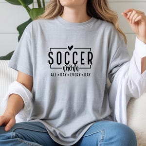 May include: A light gray t-shirt with the words "SOCCER mom" in a black font, with a heart above the word "SOCCER". The text "ALL DAY EVERY DAY" is below the words "SOCCER mom".