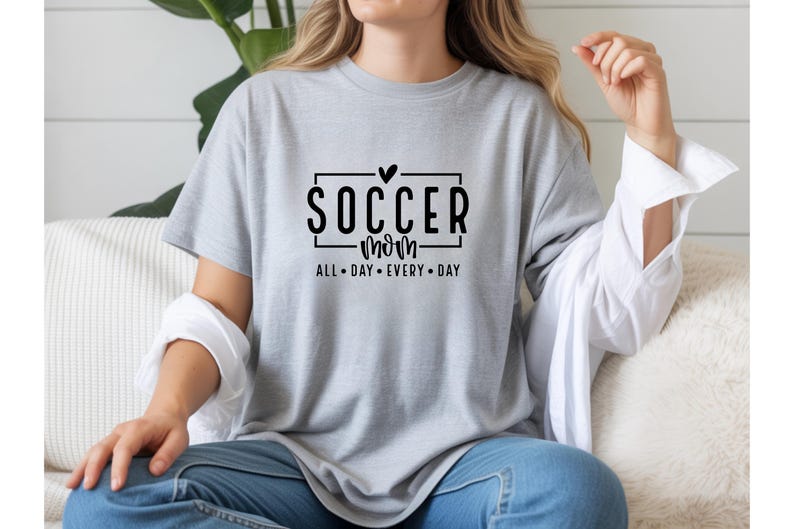 May include: A light gray t-shirt with the words "SOCCER mom" in a black font, with a heart above the word "SOCCER". The text "ALL DAY EVERY DAY" is below the words "SOCCER mom".