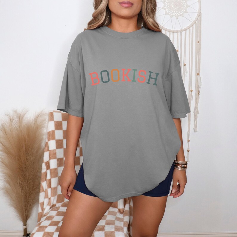 A woman wearing a gray t-shirt with the word "BOOKISH" printed on it, standing in front of a wall with a dreamcatcher.