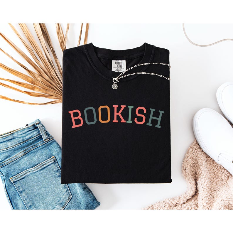 a black t-shirt with the word "BOOKISH" printed on it, along with a pair of blue jeans and white sneakers, all laid out on a white background.