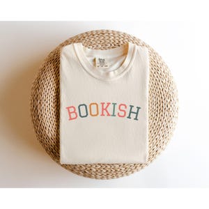 A white t-shirt with the word "BOOKISH" printed on it, laid out on a woven basket or mat.