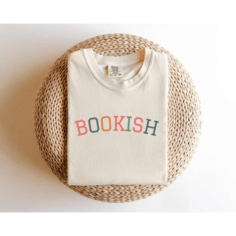 A white t-shirt with the word "BOOKISH" printed on it, laid out on a woven basket or mat.