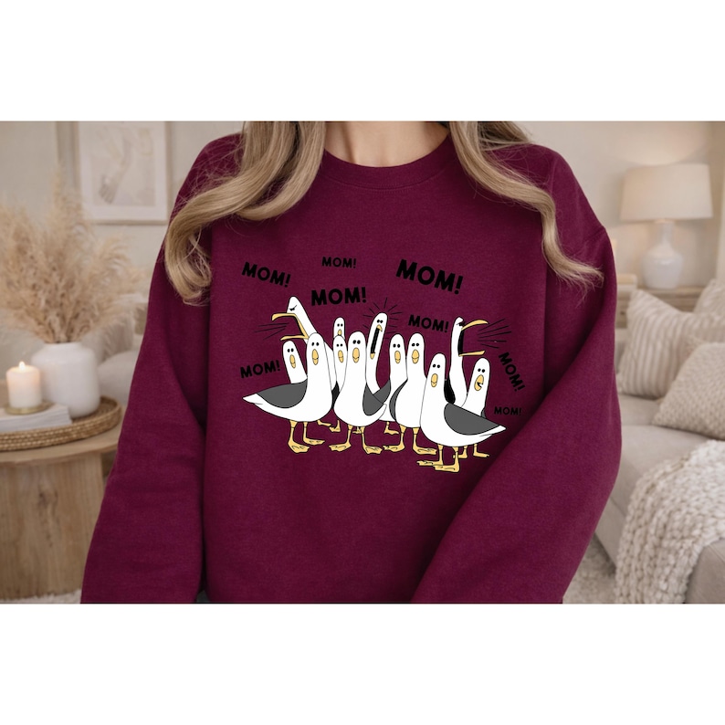 Peut inclure: Sweat-shirt bordeaux avec un motif de mouettes de dessin anim&eacute;, chacune avec le mot "MOM!" imprim&eacute; autour d'elles. Les mouettes sont blanches avec des ailes noires et des pieds jaunes. Le texte "MOM!" est en gras noir.