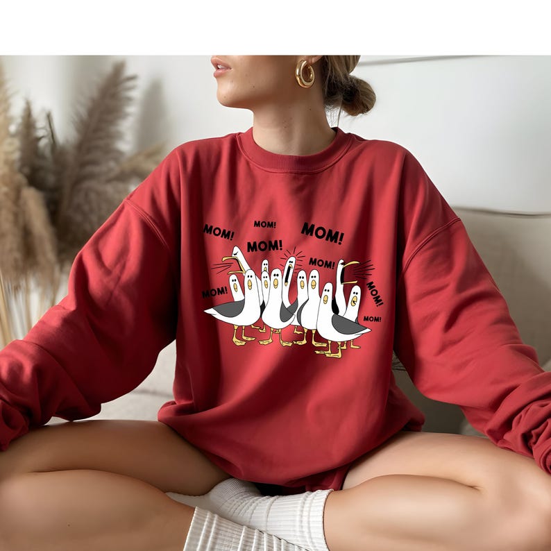 Peut inclure: Un sweat-shirt rouge avec un motif de dessin anim&eacute; de mouettes et le mot "MOM!" imprim&eacute; autour d'elles. Les mouettes sont blanches et grises avec des pieds et des becs jaunes. Le sweat-shirt est de couleur rouge uni.