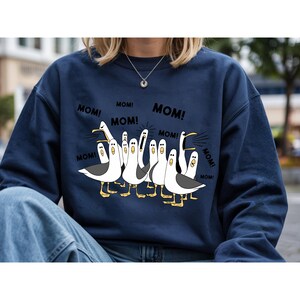 Peut inclure: Sweat-shirt bleu marine avec une illustration de mouettes criant le mot "MOM!". Les mouettes sont blanches avec des ailes grises et des pieds jaunes. Le texte "MOM!" est en noir.