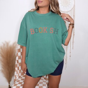 A woman wearing a green t-shirt with the word "BOOKSH" printed on it, standing in front of a dreamcatcher.