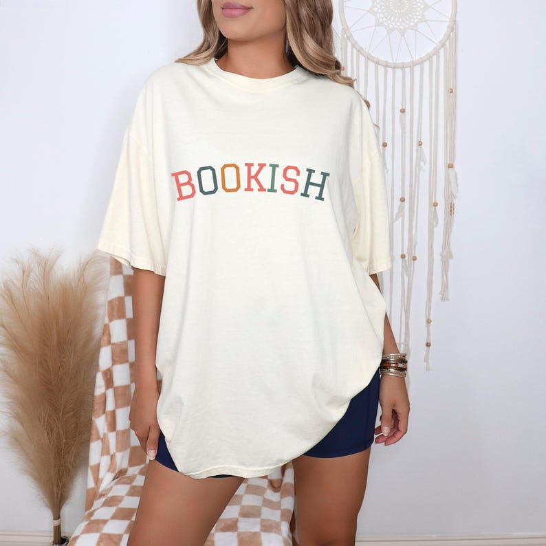 A woman wearing a white t-shirt with the word "BOOKISH" printed on it, standing in front of a dreamcatcher.