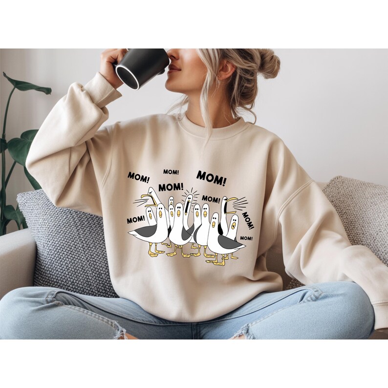 Peut inclure: Sweat-shirt beige avec un motif de mouettes criant le mot "MOM!". Les mouettes sont noires, blanches et jaunes. Le sweat-shirt est de couleur neutre et a un col rond.