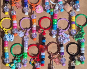 May include: A collection of colorful beaded keychains with various charms, including a bee, flower, star, and heart. Each keychain features a metal ring and a strand of beads in a rainbow of colors. The charms add a playful touch to the accessories.