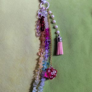 Könnte beinhalten: Ein handgefertigter Schlüsselanhänger mit Karabinerverschluss, der eine Vielzahl farbiger Perlen, eine goldene Kette, Kunstperlen und eine rosa Quaste aufweist. Ein roter, durchscheinender, 12-seitiger Würfel ist an der Kette befestigt.