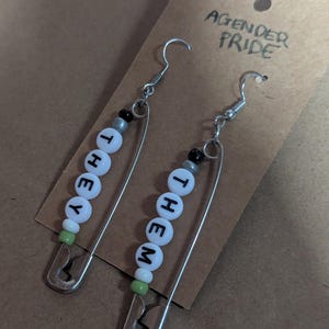 May include: A pair of handmade earrings featuring silver-tone safety pins. Each earring has white letter beads spelling "THEY" and "THEM", with additional black, gray, and green beads. The earrings are displayed on a brown card with "AGENDER PRIDE" written on it.