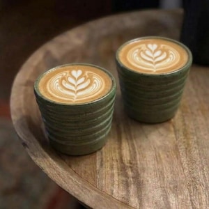 May include: Two ceramic cups with a ribbed, olive-green speckled finish, each containing a coffee beverage topped with a white foam heart-shaped latte art design. The cups are cylindrical with horizontal ridges along the exterior surface.