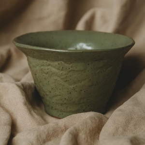 Może przedstawiać: Ręcznie wykonana, oliwkowozielona ceramiczna miska o teksturowanej powierzchni zewnętrznej i lekko rozszerzonym brzegu. Miska ma nakrapiany wygląd i stoi na tkaninie o neutralnym odcieniu, prezentując swoje ziemiste odcienie i rzemieślniczą jakość.