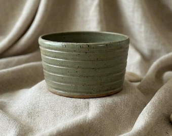 May include: A sage green ceramic bowl with a speckled finish and horizontal ribbed texture along the exterior walls. The bowl features a wide, open circular rim and a smooth, rounded base, showcasing a consistent glaze throughout the piece.