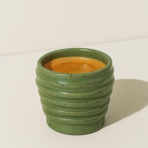 Puede incluir: Una pequeña taza de cerámica verde con un exterior texturizado y acanalado. La taza está llena de un líquido marrón claro, posiblemente café o espresso. La taza está moteada y se encuentra sobre una superficie blanca.