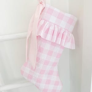 Pode incluir: Uma meia de Natal acolchoada com um padrão de xadrez vichy em rosa claro e branco. A meia inclui um punho com folhos na parte superior e uma fita comprida de cor rosa claro sólido atada num laço na alça de suspensão decorativa.