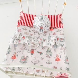 May include: A miniature white metal bed frame featuring a headboard with gold-toned finials. The bedding includes a white pillow with red vertical stripes, a round fabric accent, and a white quilt patterned with Christmas-themed illustrations.