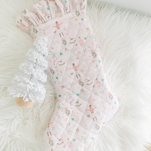 May include: A quilted Christmas stocking in a pale pink fabric featuring a repeating pattern of nutcrackers, rocking horses, and greenery. The stocking has a ruffled top edge and is accompanied by a small white textured decorative tree.