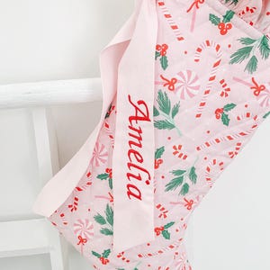 Puede incluir: Un calcetín navideño acolchado de color rosa claro con un patrón repetitivo de bastones de caramelo rojos y blancos, ramas de pino verdes y bayas de acebo rojas. Incluye una cinta rosa claro bordada con el nombre Amelia en letra roja.