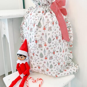 May include: A white fabric gift sack featuring a repeating pattern of Christmas-themed illustrations including nutcrackers, ballerinas, and trees, finished with a red and white striped ribbon bow and ruffled side edges, accompanied by a red elf doll.