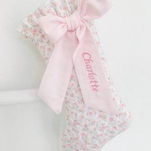 May include: A quilted Christmas stocking featuring a white fabric with a small pink floral pattern. The stocking has a ruffled top edge and a large, solid light pink ribbon bow attached to the front. The name Charlotte is embroidered in pink.