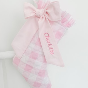 Peut inclure: Une chaussette de Noël matelassée avec un motif vichy rose clair et blanc. La chaussette est ornée d'un grand nœud en tissu rose clair uni, et le prénom Charlotte est brodé en fil rose sur l'un des longs pans du nœud.