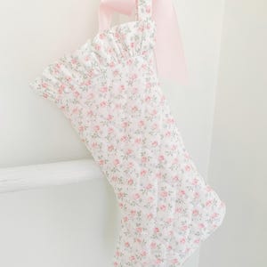 Pode incluir: Uma meia de Natal acolchoada com base de tecido branco e um pequeno padrão floral rosa. A meia tem uma borda superior com babados e uma fita rosa claro amarrada em um laço na alça de suspensão, proporcionando um visual suave e decorativo.