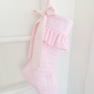 May include: A quilted Christmas stocking made of light pink and white striped fabric. The stocking features a ruffled cuff and a solid light pink ribbon bow tied at the top. It includes a matching striped fabric loop for hanging on a white wooden post.