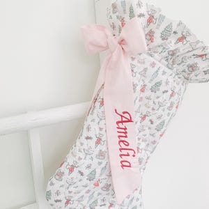 Pode incluir: Uma meia de Natal acolchoada em tecido branco com um padrão repetitivo de pequenas figuras, árvores e presentes em vermelho, verde e azul. Uma fita rosa claro está atada num laço no topo, bordada com o nome Amelia em vermelho.